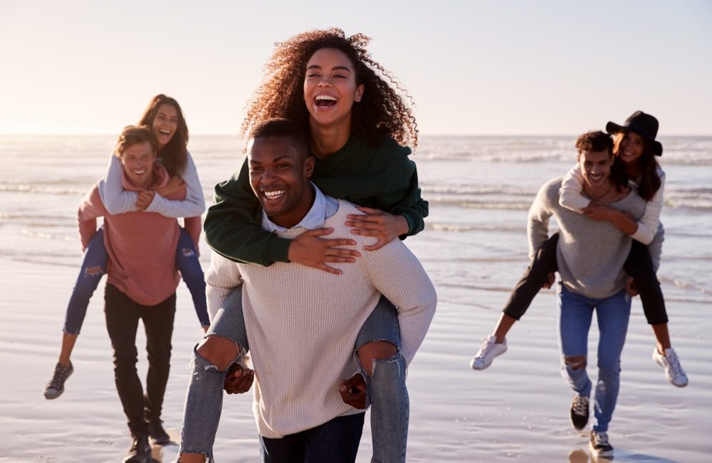 Group,Of,Friends,Having,Piggyback,Race,On,Winter,Beach,Together